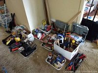 Overview of multiple boxes containing assorted hand tools, toolkits, hardware, flashlights, and painting supplies arranged on carpeted floor