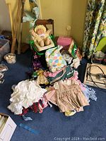 Cabbage Patch Kid doll seated in wooden child's chair with green plastic frame, surrounded by vintage baby clothes, international dolls and vintage books on the floor.