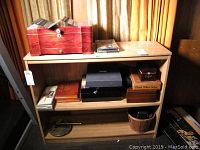 Shelf holding several cigar humidors and boxes, metal cutters, a basket, and a VHS tape, all related to cigar storage and maintenance.