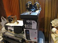 Photo showing TrippLite Battery Backup UPS tower next to AIWA cassette radio and VHS player with several headphones resting on top.
