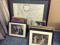 Framed assortment of art pieces including prints, needlepoint, and a vintage map arranged on carpet in front of curtain and wall.