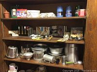 Wooden shelving unit holding a variety of metal kitchenware items including pans, molds, grinders, and other utensils along with packaged beeswax and other miscellaneous items.