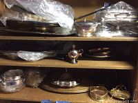 Multiple silver plated trays, chafing dish and other serving items shown on shelves, some wrapped in plastic.