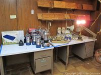 Wide view of table with Coleman and GE lanterns, brass lamps, and boxed air bed.