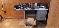Overview of multiple binoculars of various sizes arranged on table and in a box on floor, some dusty and with wear.