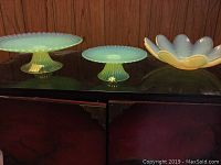 Two green glass cake stands of different sizes and one green glass decorative bowl with scalloped edges placed on a black surface against wood paneling.