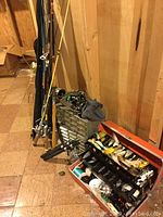 Photo shows multiple fishing rods and poles leaning against a wooden wall, with some reels attached. Below them are hardware organizers with small drawers and an open tackle box filled with colorful fly fishing lures.