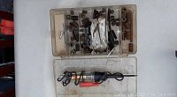 Photo showing a plastic compartment box filled with metal fittings such as springs, screws, nuts, bolts, washers, and an electric test tool with attached cables, placed on a white surface.