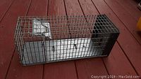 Metal wire mesh live-catch rodent trap with spring door lying on wooden floor, showing side view.