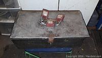 Closed metal toolbox with three red padlocks on top, showing weathering and rust.