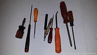 Photo shows eight various small screwdrivers and similar tools laid out horizontally on a white surface. Handles range from plastic to wood in multiple colors.