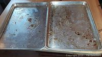 Two large metal baking trays placed side by side on a kitchen counter, showing wear and stains.