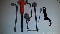 Photo showing seven assorted hand tools on white surface including wrenches, screwdriver with red handle, a crank tool, small blue cleaning brush, and a spanner wrench.