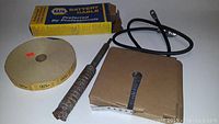 Photo showing all four items: NAPA battery cable in box, a roll of wire support, a large rusty drill bit, and a box of metal tickets or labels.
