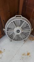 Full view of the white 24 inch fan with handle and two legs seen from the front, located on wooden floor and near the wall.