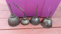 Four vintage metal oil cans lined up on a wooden surface showing different spout styles and wear.