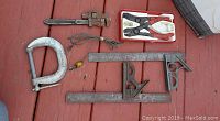 Photo showing a lot of mechanical tools including an adjustable pipe wrench, a large metal C-clamp, pliers in a plastic tray, and two bar-style woodworking clamps with attached squares.