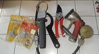 Top-down image showing a variety of hand tools arranged side by side including a clamp, sprayer nozzle, pruning clipper, pressure gauge, and packaged parts.