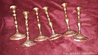 Photo showing six brass candlesticks standing upright on a dark red surface, arranged in a row.