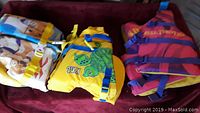Three youth flotation devices laid out on a surface: one with an animal pattern, one yellow with a turtle image, one red and yellow.