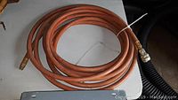 Coiled orange air pressure hose approximately 15 feet long placed on white surface. Shows metal fittings on each end.