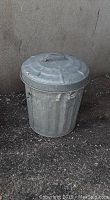 Photo of a weathered metal trash can about 2 feet tall with a domed lid and handle, sitting outdoors on gravel against a concrete wall.