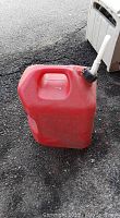 Photo of a red 5 gallon plastic gas can with attached white spout, placed on asphalt surface near a grey container.