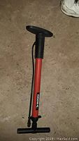 Photo of a red and black Husky manual bicycle pump with hose and T-handle, standing on a rough floor surface.