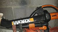 Orange and black WORX electric leaf blower resting against a wall, with visible power cord and handle.