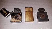 Photo showing three vintage lighters, one with gold-tone finish, one silver-tone with emblem, one silver-tone with graphic image and text, all open or partially open.