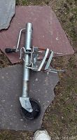 Metal boat trolley lift assembly lying on stone slabs and grass, showing single black wheel, handle, springs, and mounting hardware.