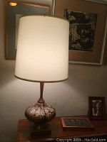 Full view of the lit lamp showing the off-white fabric shade and overall height on teak furniture.