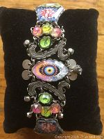 Full view of bracelet showing central oval cabochon with eye-like pattern, flanked by green and pink bead clusters and decorative silver-tone metal scrolling.