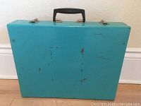 Front view of the turquoise metal suitcase showing scratches and wear.