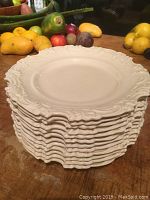 Stack of 12 buttery cream colored Royal Doulton plates with ornate decorative edges shown stacked on wooden surface with fruit in background.