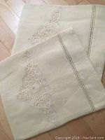 Two cream linen pillow shams with embroidered designs and lace cutwork folds, laid flat on wooden floor.