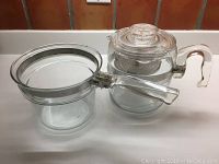 Two clear glass kitchenware items including a coffee percolator and a glass pot with handle on white surface
