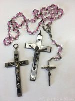 Three Christian crosses including two decorative crosses and a pink beaded rosary on a white background.