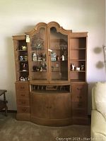 Front view of wooden china cabinet showing scalloped top, glass doors with locking mechanism, side wooden shelves with decorative items, drawer handles, and bottom cabinet with curved doors.