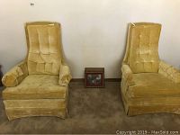 Both vintage yellow upholstered high back chairs side by side against a white wall on carpeted floor. Wooden framed photo or artwork on the floor between chairs.