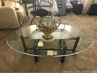 Oval glass coffee table with wood base shown from side angle with decorative vase on top (vase not included in sale).