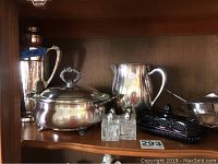 Shelf with tall silver pitcher, silver butter dish, glass salt and pepper shakers, black glass covered dish, and silver serving bowl with ladle.