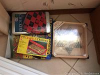 Box containing Checkers tin, Double 6 Dominoes box, and dollhouse wall decor frame with small silver bells.
