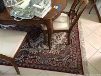 First large area rug underneath a table and chairs showing floral and border patterns in cream and red tones with fringe