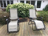 Pair of two tan zero gravity lounge chairs placed on a wooden deck in front of a white house with green shrubbery behind.