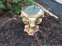 Concrete sundial base with four lion heads and paws facing outward supporting an octagonal top with visible wear and moss.
