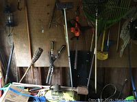 Greenworks pressure washer with hose, Black and Decker hedge trimmer, garden rakes, tiki torch, pruners, and other hand tools hanging on pegboard and leaning against wall.