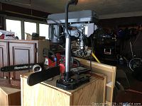 Side view of the Central Machinery drill press mounted on a wooden workbench with surrounding workshop items visible, showing its upper motor and drill head.
