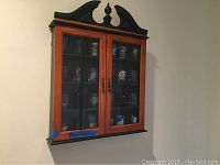 Wall-mounted wooden cabinet painted dark green with natural wood-framed glass doors and ornate top piece, closed on wall