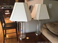 Two matching table lamps with clear glass bases and white cone-shaped shades, positioned on a wooden table near a sofa and chairs.